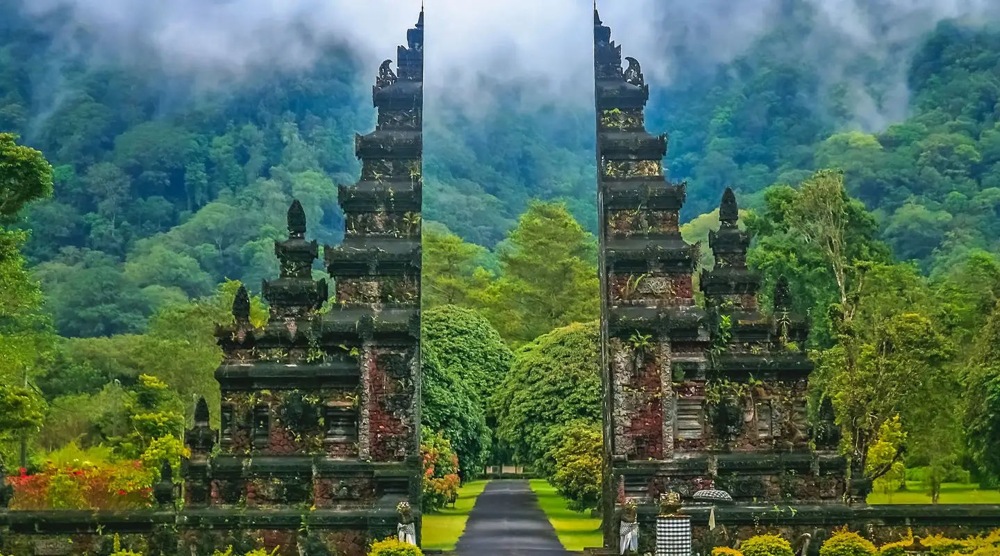Puerta del templo Handara en Bali, Indonesia, con montañas verdes y niebla de fondo, rodeada de exuberante vegetación.