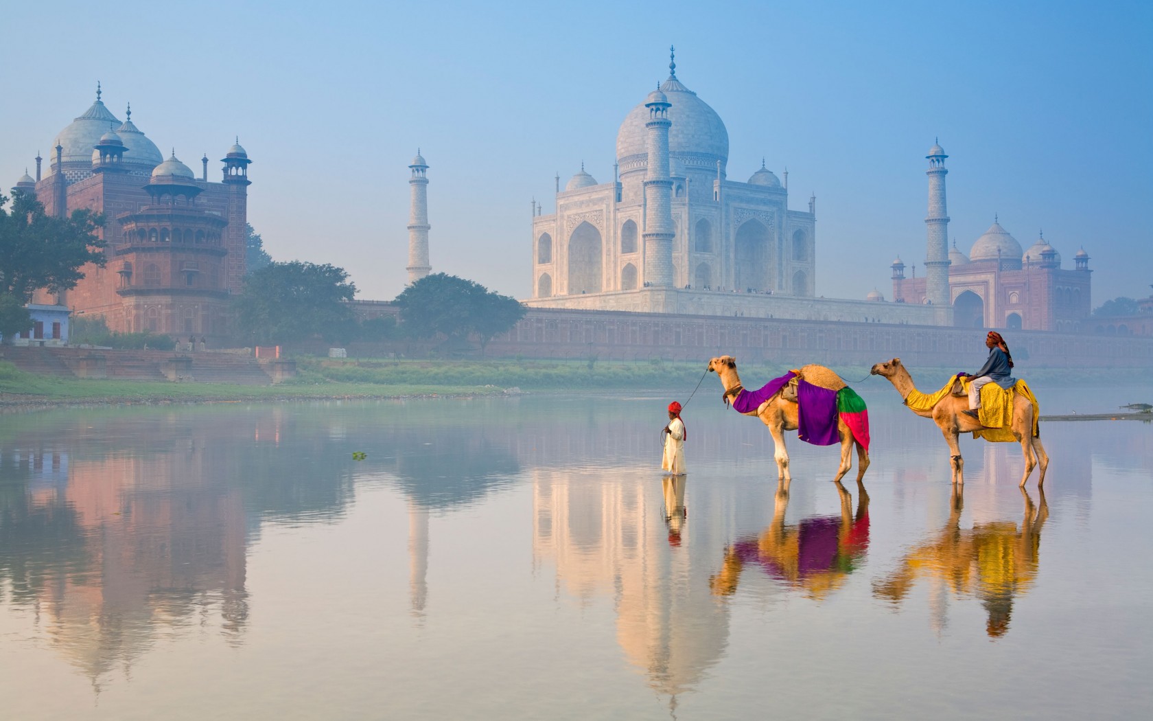 Dos hombres con camellos cruzando un río con el Taj Mahal de fondo.