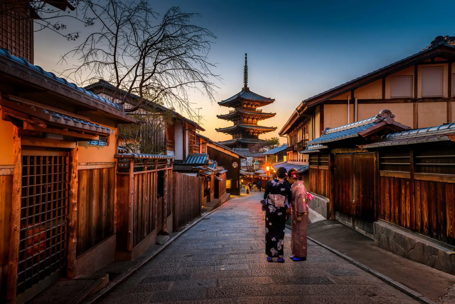 Calle tradicional en Kyoto, Japón, con casas de madera, un templo pagoda al fondo y dos personas vistiendo kimonos al atardecer.
